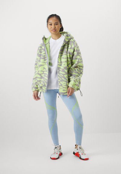 Casaco de fleece com capuz em padrões verdes e cinzentos, combinado com leggings azul claro com acentos em neon e ténis brancos.