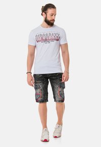 Weißes T-Shirt mit grafischem Druck und schwarze Cargo-Shorts mit Reißverschlusstaschen. Die Schuhe sind weiß-rote Sneakers.