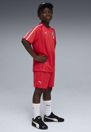 Garçon portant un équipement de football rouge, une casquette noire, des chaussettes blanches Puma et des baskets noires Puma, debout les mains sur les hanches devant un fond gris.