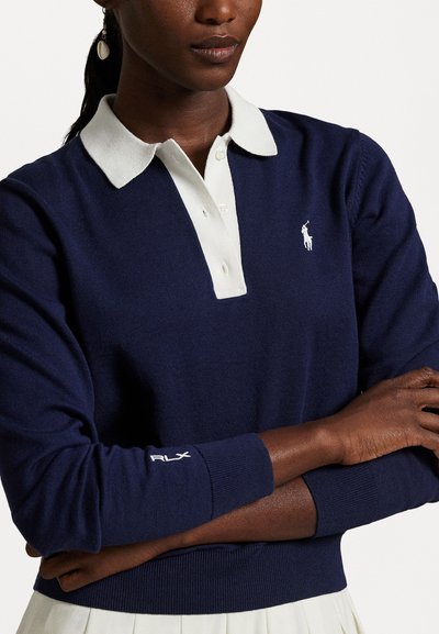 Pull en maille bleu marine avec un col polo blanc, présentant un petit logo polo sur la poitrine et un broderie "RLX" sur la manche.