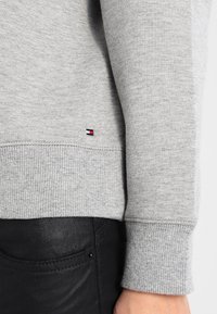 Szary bawełniany sweatshirt z prążkowanymi mankietami. Ma małe logo na dolnym brzegu. W połączeniu z czarnymi spodniami, ukazującymi teksturowany materiał.