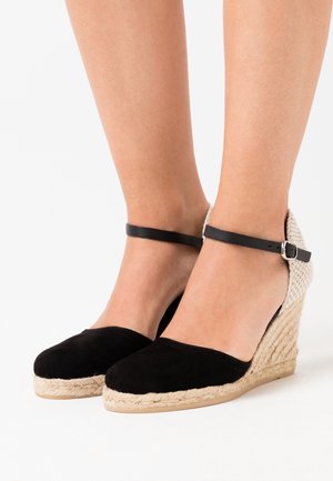 Des jambes portant des espadrilles compensées noires avec des semelles en jute tressé et des brides autour des chevilles, se tenant sur un fond blanc.