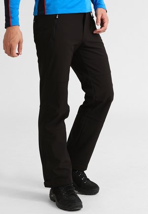 Mand iført sorte trekkingbukser med lynlåslommer og sorte vandrestøvler, stående foran en ensfarvet baggrund.