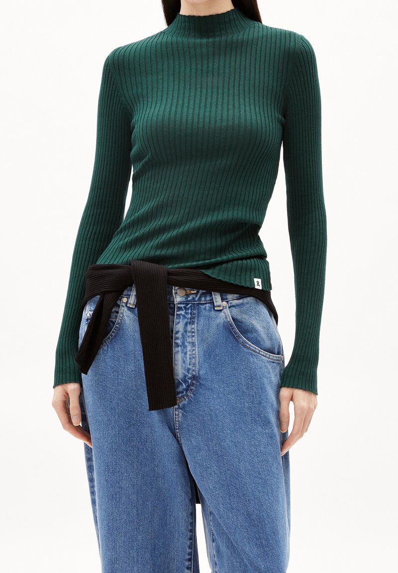 Maglia verde a costine con maniche lunghe e collo alto, impreziosita da un nastro nero in vita, abbinata a jeans a gamba larga blu chiaro.