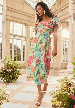 Frau in einem geblümten, pastellfarbenen Kleid und goldenen Absätzen, die drinnen zwischen Blumenarrangements in einem hellen, geräumigen Raum mit großen Fenstern geht.