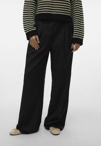 Pantalons amples noirs avec plis sur le devant, associés à un pull en maille rayé beige et noir, et des ballerines beiges avec des nœuds.