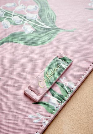 Rózsaszín, texturált szőnyeg virágos mintával, zöld levelekkel és fehér virágokkal; tartalmaz egy téglalap alakú címkét arany színnel, melyen a "Cath Kidston" felirat szerepel.