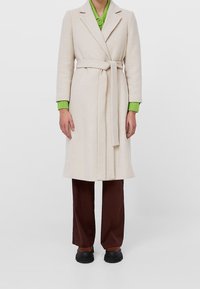 Manteau long beige avec une ceinture, fabriqué en tissu doux. Avec des revers crantés, détail de couture d'épaule, porté sur un pull vert et un pantalon marron. Chaussures noires.