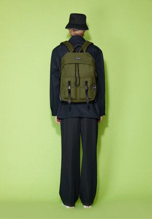 Persona de espaldas, con un sombrero tipo bucket negro, ropa oscura y una mochila verde oliva, frente a un fondo verde claro.