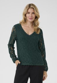 Top in pizzo verde scuro con maniche lunghe, scollatura a V con bordo ondulato e motivo floreale testurizzato. Il materiale è delicato con un effetto trasparente sulle maniche.