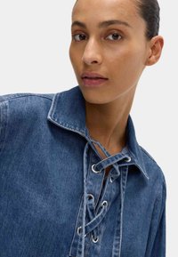 Chemise en denim avec un décolleté à lacets, des œillets en métal argenté et un col pointu. Le tissu est bleu moyen et a une texture lisse.