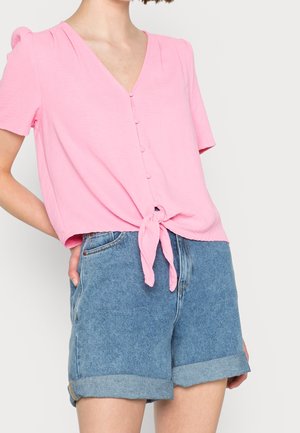 Femme portant un chemisier rose clair à manches courtes avec des boutons et un nœud à l'avant, associé à un short en jean bleu avec un ourlet retroussé.
