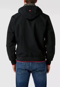 Veste noire à capuche avec des poignets et un ourlet élastiques, présentant un accent rouge contrastant en bas. Doublure à carreaux grise visible au niveau du col.