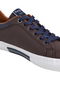 Barna bőrből készült sportcipő tengerészkék fűzőkkel, fehér gumitalppal és világosbarna béléssel. A nyelvén logócímke és texturált oldalpanelek találhatók.