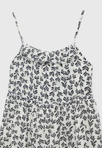 Robe blanche sans manches avec fines bretelles et motif de feuilles noires, avec un col à volants et une taille froncée.