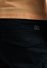 Pantaloni neri con una tasca posteriore a destra, caratterizzata da dettagli cuciti e una piccola etichetta rettangolare del marchio. Orlo arancione della maglietta visibile sopra.