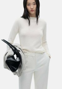Maglione color crema con una consistenza morbida, abbinato a pantaloni bianchi e una sciarpa grigia appoggiata su un braccio. Borsa nera tenuta nell'altra mano.