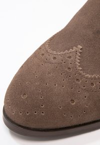 Bout de chaussure en daim marron avec des perforations décoratives et un détail de surpiqûres festonnées sur une semelle foncée.