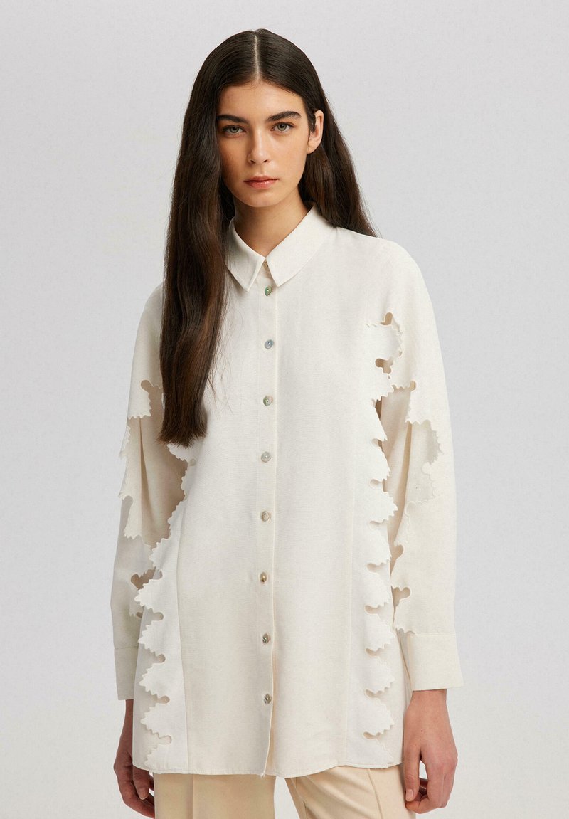 Camicia beige chiaro con colletto aderente, bottoni sul davanti e motivi a foglia ritagliati lungo le maniche. Tessuto di consistenza liscia.
