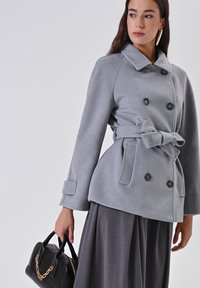 Cappotto grigio doppiopetto con cintura, caratterizzato da un tessuto testurizzato e grandi bottoni, abbinato a una gonna grigio scuro e una borsa nera.