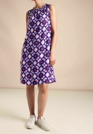 Sleeveless purple dress with geometric pattern, worn by standing person in white sneakers against plain beige background.