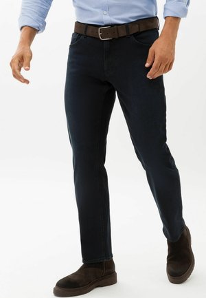 Mand iført en lyseblå skjorte, mørke slim-fit jeans med brunt bælte og mørkebrune ruskindsstøvler, stående mod en hvid baggrund.