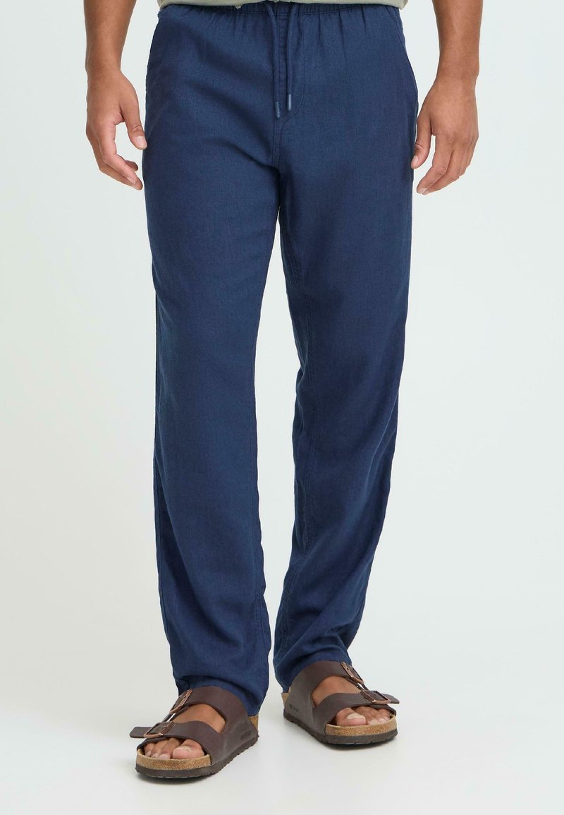 Pantaloni ușori din in de culoare albastru marin, cu talie elastică și șnur, asortați cu sandale maro cu cataramă, având tălpi texturate.