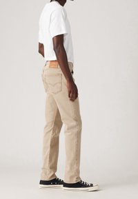 Jeans beige de corte recto con bolsillos traseros, combinados con una camiseta blanca y zapatillas negras. Textura desgastada y parche de cuero de marca.