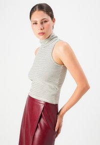 Mladá žena s vlasy zviazanými dozadu, oblečená v tielku s prúžkami a vysokým pásom červenej koženkovej sukne na bielom pozadí.