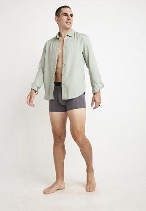 Lysegrøn skjorte med knapper, korte ærmer, åben ved brystet, båret over mørkegrå boxer-shorts. Modellen står barfodet imod en neutral baggrund.