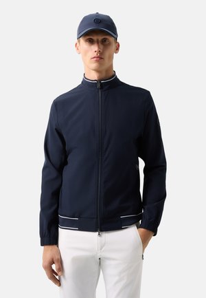 Homme portant une veste zippée marine avec des détails en bandes blanches, un pantalon blanc et une casquette de baseball marine, debout devant un fond uni.