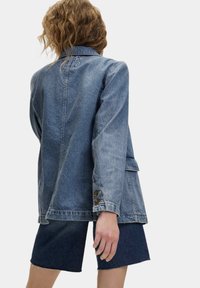 Giacca di jeans azzurro chiaro con una vestibilità ampia, dotata di colletto a punta, polsini con bottone e tasche laterali. Abbinata a pantaloni corti di jeans scuri.