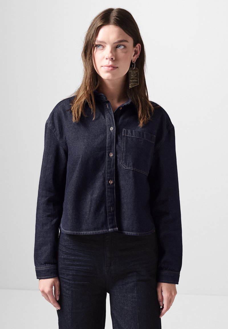 Camicetta corta in denim scuro con colletto a punta, tasca frontale e chiusura a bottoni. Tessuto testurizzato con dettagli di cucitura in contrasto.