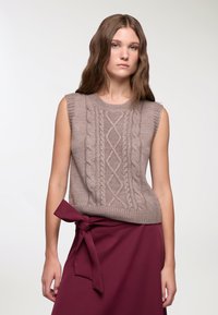 Beżowy bezrękawnik z dzianiny, zdobiony wzorem w warkocze, z prążkowanym dekoltem i dołem. Noszony z bordową spódnicą z wiązanym paskiem.