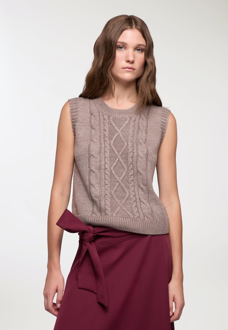Beżowy bezrękawnik z dzianiny, zdobiony wzorem w warkocze, z prążkowanym dekoltem i dołem. Noszony z bordową spódnicą z wiązanym paskiem.
