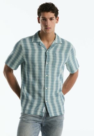 Junger Mann mit lockigem Haar, trägt ein kurzärmeliges blau-weiß gestreiftes Hemd mit Knöpfen und hellblaue Jeans, mit den Händen hinter dem Rücken.