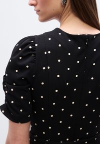 Γυναίκα με μαύρο φόρεμα με λευκές πουά, φουσκωτά κοντά μανίκια και ασημένια σκουλαρίκια κρίκους, εμφανιζόμενη από την πλάτη και το πλ άι.