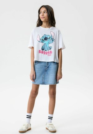 Ragazza che indossa una maglietta bianca oversize con il cartone di Stitch, gonna di jeans, calzini bianchi con strisce nere e scarpe da ginnastica bianche, in piedi davanti a uno sfondo neutro.