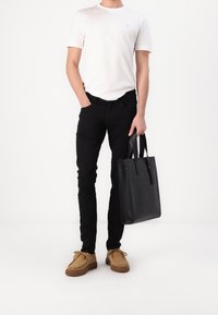 Bolso tote de cuero negro con dos asas, llevado por una persona con una camiseta blanca y pantalones negros, de pie sobre un fondo blanco.