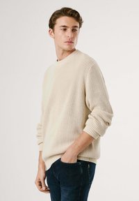 Cremfarbener, gerippter Pullover mit hohem Kragen und umgeschlagenen Ärmeln, getragen über dunkelblauen Jeans. Strickstruktur sichtbar.