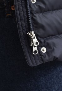 Tamsiai mėlynas pūkinis švarkas su dvikrypčiu užtrauktuku ir metalinėmis spaudėmis, su siuvinėta tekstūra ir lygiu audiniu, fone tamsaus džinso.