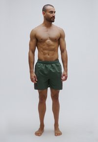 Πράσινα μαγιό σορτς με ελαστική μέση, με διακριτή υφή και πλαϊνές τσέπες, φορεμένα από έναν άνδρα μοντέλο που στέκεται ξυπόλητος.