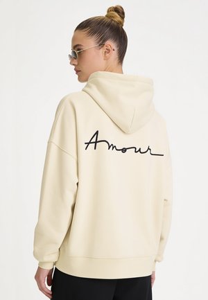 Femme avec les cheveux en chignon, portant des lunettes de soleil et un sweat à capuche crème avec un texte noir "Amour" dans le dos, de profil sur fond blanc.