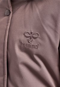 Chemise en tissu léger marron avec une texture lisse, ornée d'un logo brodé dans une teinte plus foncée. Fermée par des boutons moulés.