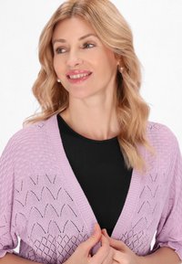 Cardigan a maglia lilla con apertura frontale, motivo a rombi testurizzato e maniche corte sopra una maglietta nera, indossato da una persona con capelli chiari.