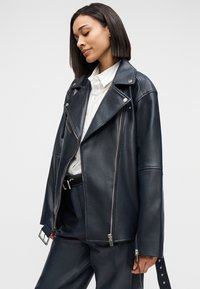 Femme aux cheveux mi-longs portant une veste en cuir noire, une chemise blanche à col, un collier de perles et un pantalon en cuir noir, debout les yeux fermés.