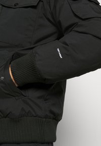 Personne portant une veste noire avec des poignets et une ceinture côtelés, main dans la poche, et texte blanc "DRYVENT" sur la manche.