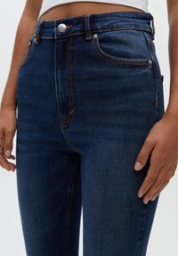 Höga, mörkblå denimjeans med orange sömmar och knappstängning. Har framfickor och en slät yta.