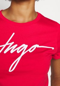 Camiseta de algodón roja con un destacado logo en blanco en escritura "Hugo" en el frente. Se pueden ver las mangas cortas y un escote redondo.