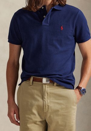 Homme portant un polo bleu marine avec un logo rouge, un pantalon beige, une ceinture marron et une montre-bracelet bleue avec un bracelet en cuir marron.